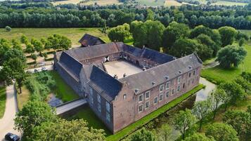 Een groot, historisch gebouw met een binnenplaats, omgeven door weelderige begroeiing en velden.