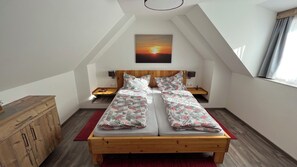 A bedroom with a sloped ceiling, a bed with a wooden frame, two bedside tables, a nightstand, a red rug, and a painting of a sunset. - Holzer Vacation Home (Mönichwald)
