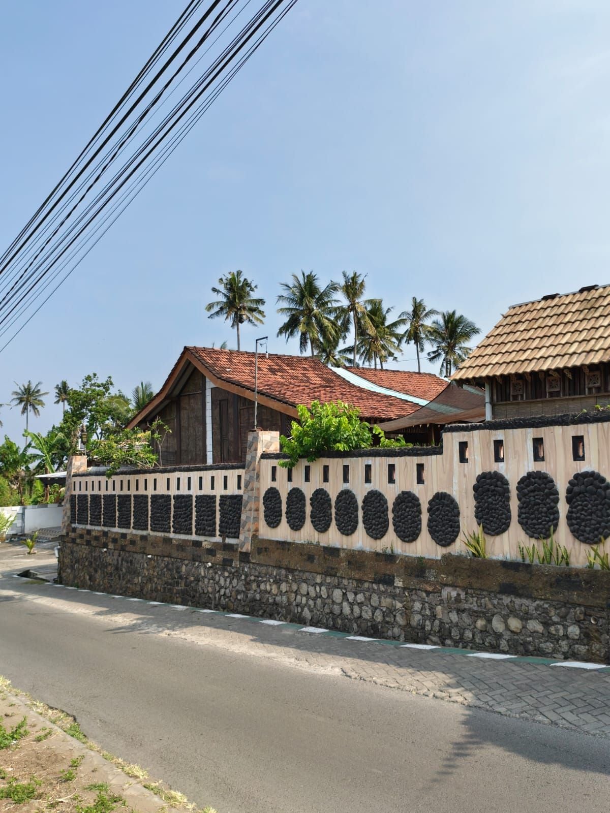 A residential area with a stone wall, a wooden house with a tiled roof, and palm trees in the background.