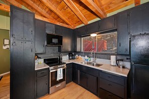 A compact kitchen with dark cabinets, a stainless steel stove, and a window with a view of a red building. - Lake View Cabin Game Room (Crestline)