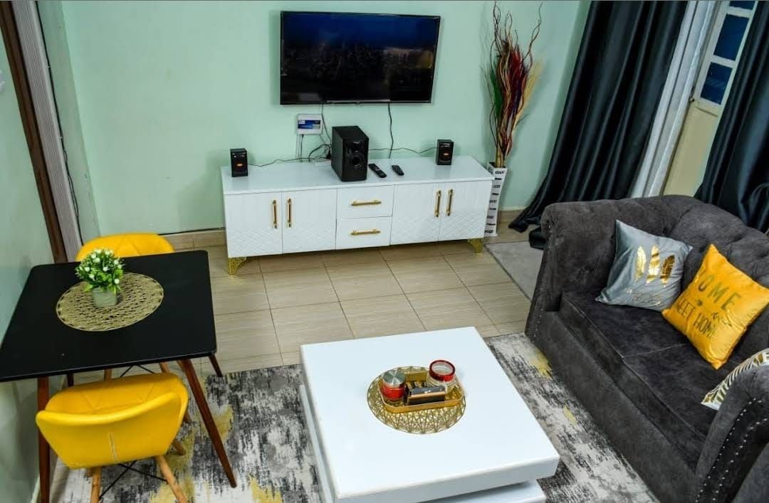 A living room with a white cabinet, a black coffee table, a grey sofa with a yellow pillow, and a television mounted on the wall.