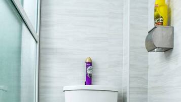 Un baño con un inodoro blanco, un cubo de basura verde y un estante en la pared con productos de limpieza.