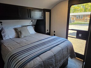A bed with a striped blanket, a headboard, and a window with a view of a building and trees. - Russells River Retreat (Gadsden)
