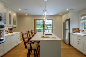 A modern kitchen with white cabinetry, a central island, wooden chairs, a stainless steel refrigerator, and a large window with a view of greenery. - Wooded Retreat w/ Porch & Fire Pit in Stone Ridge (Stone Ridge)