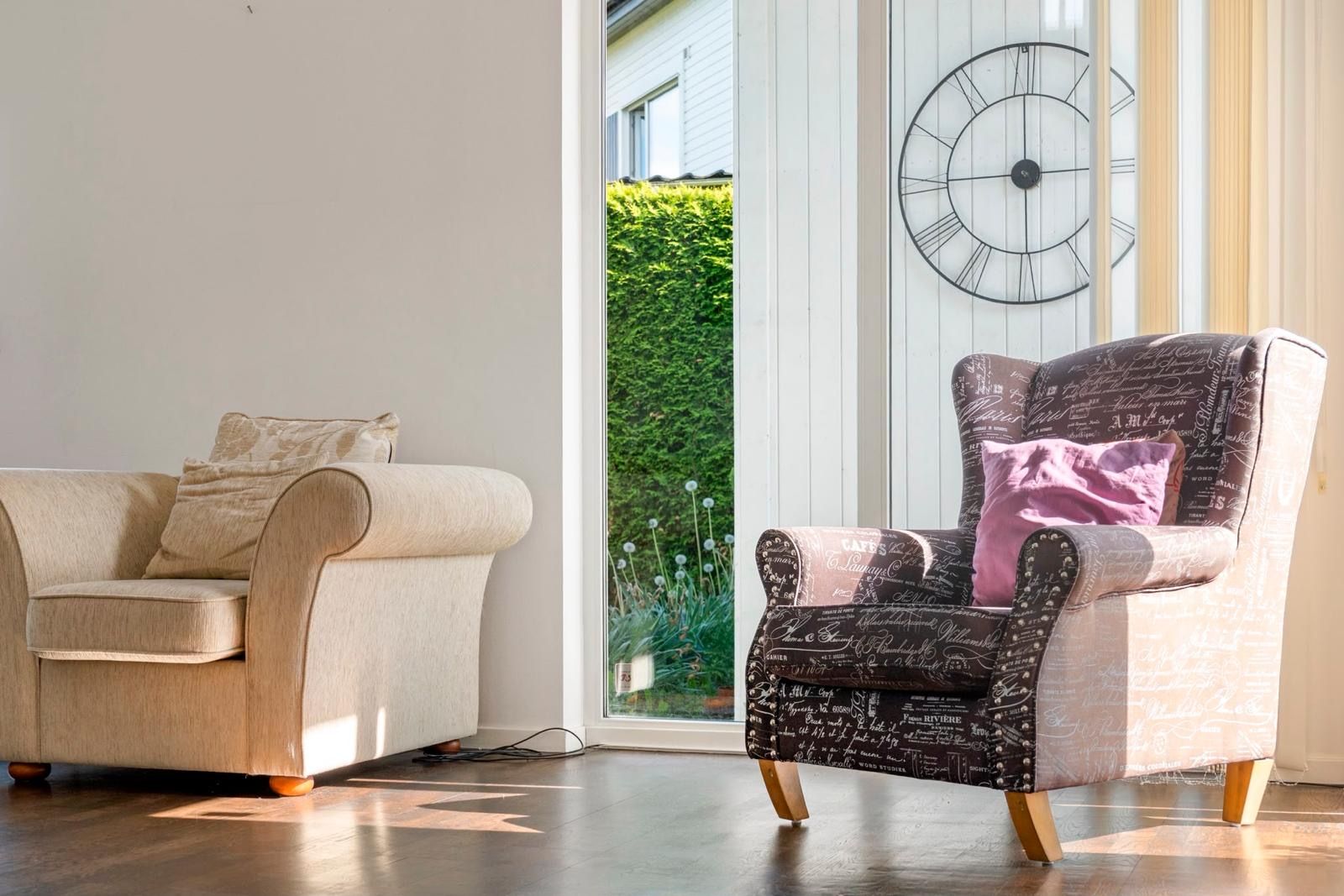 Un salon avec un canapé et un fauteuil, une grande fenêtre donnant sur un paysage verdoyant et une horloge murale.