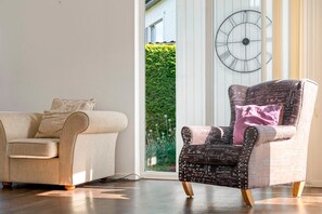 A living room with a sofa and a chair, a large window with a view of greenery, and a wall clock. - Charming Bro Retreat, Luxury Comfort & Modern Amenities (Bro)