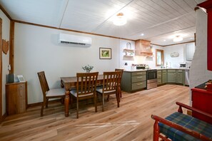 A dining area with wooden furniture, a kitchen with green cabinets, and an air conditioning unit on the wall. - Hocking Hills Area Cabin | Walk to National Forest (Nelsonville)