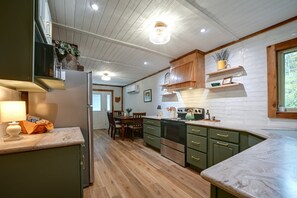 A kitchen with wooden cabinets, a marble countertop, and a dining area with a table and chairs. - Hocking Hills Area Cabin | Walk to National Forest (Nelsonville)