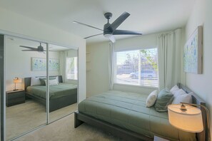 Een slaapkamer met een bed, een plafondventilator, een raam met jaloezieën en een spiegel.