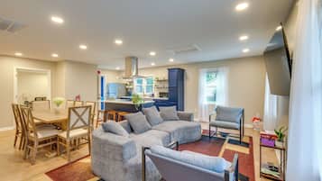 A modern living room with a sectional sofa, a dining table with chairs, a kitchen area, and a large window with curtains.
