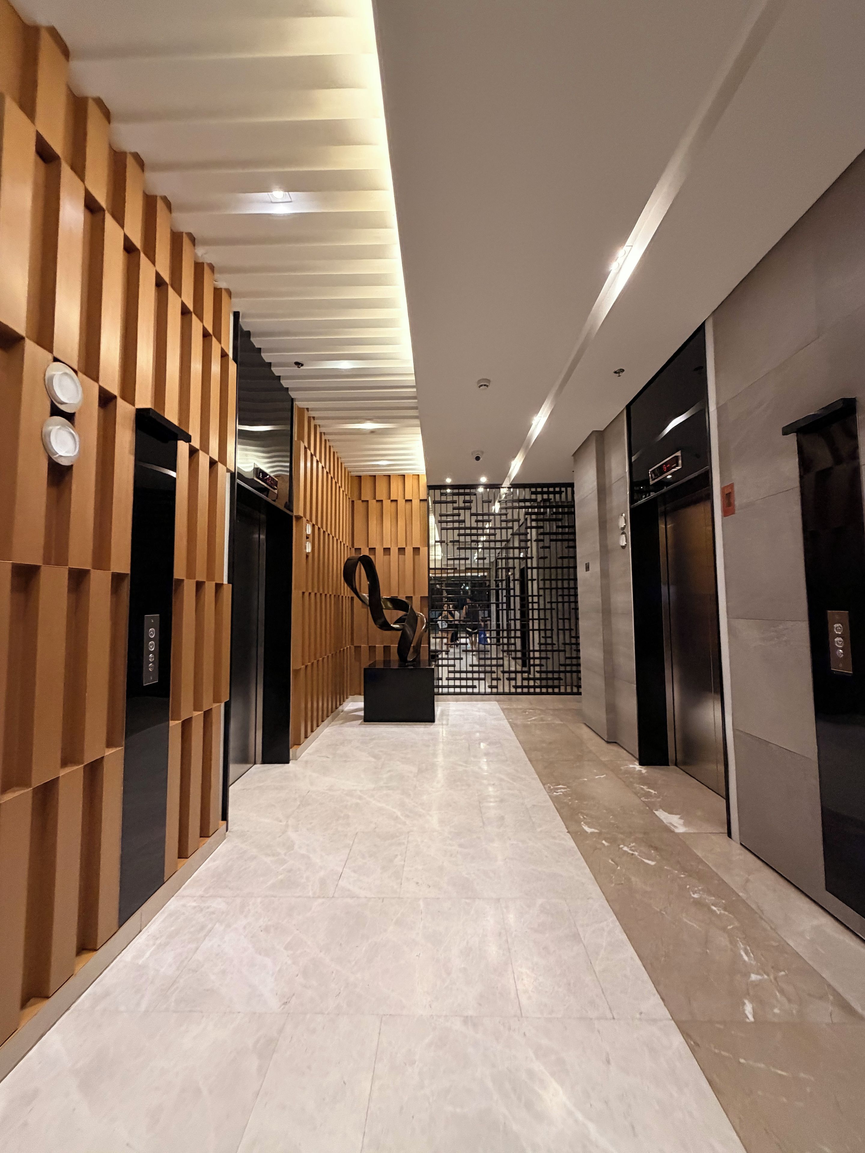 A modern interior with a hallway leading to elevators, featuring a wooden panel wall and a marble floor.