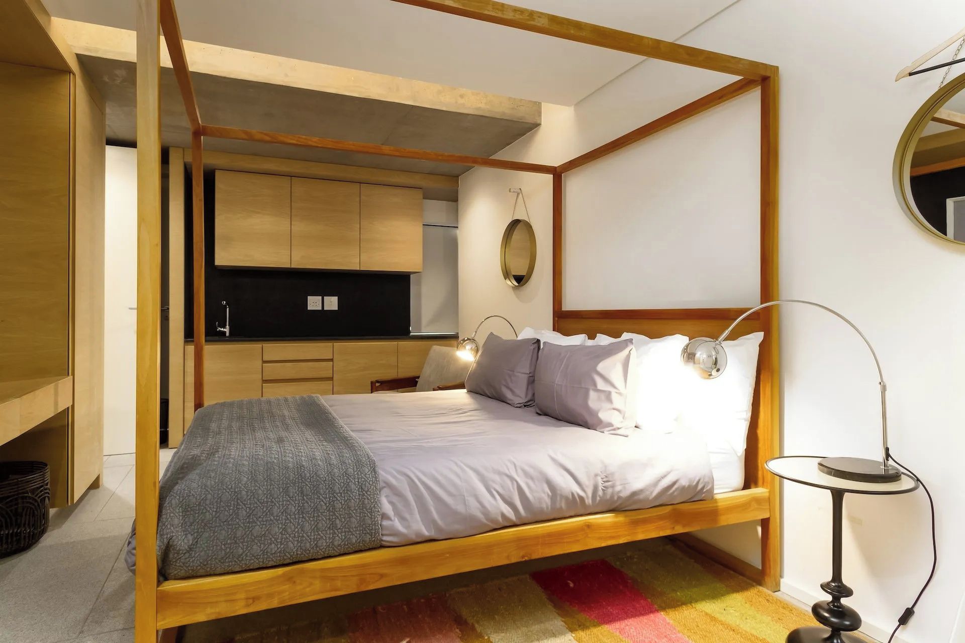 Une chambre à coucher avec un lit à baldaquin en bois, une table de chevet avec une lampe, un petit miroir rond et un coin cuisine équipé de placards et d’un plan de travail.