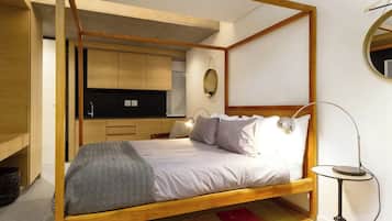 Une chambre à coucher avec un lit à baldaquin en bois, une table de chevet avec une lampe, un petit miroir rond et un coin cuisine équipé de placards et d’un plan de travail.