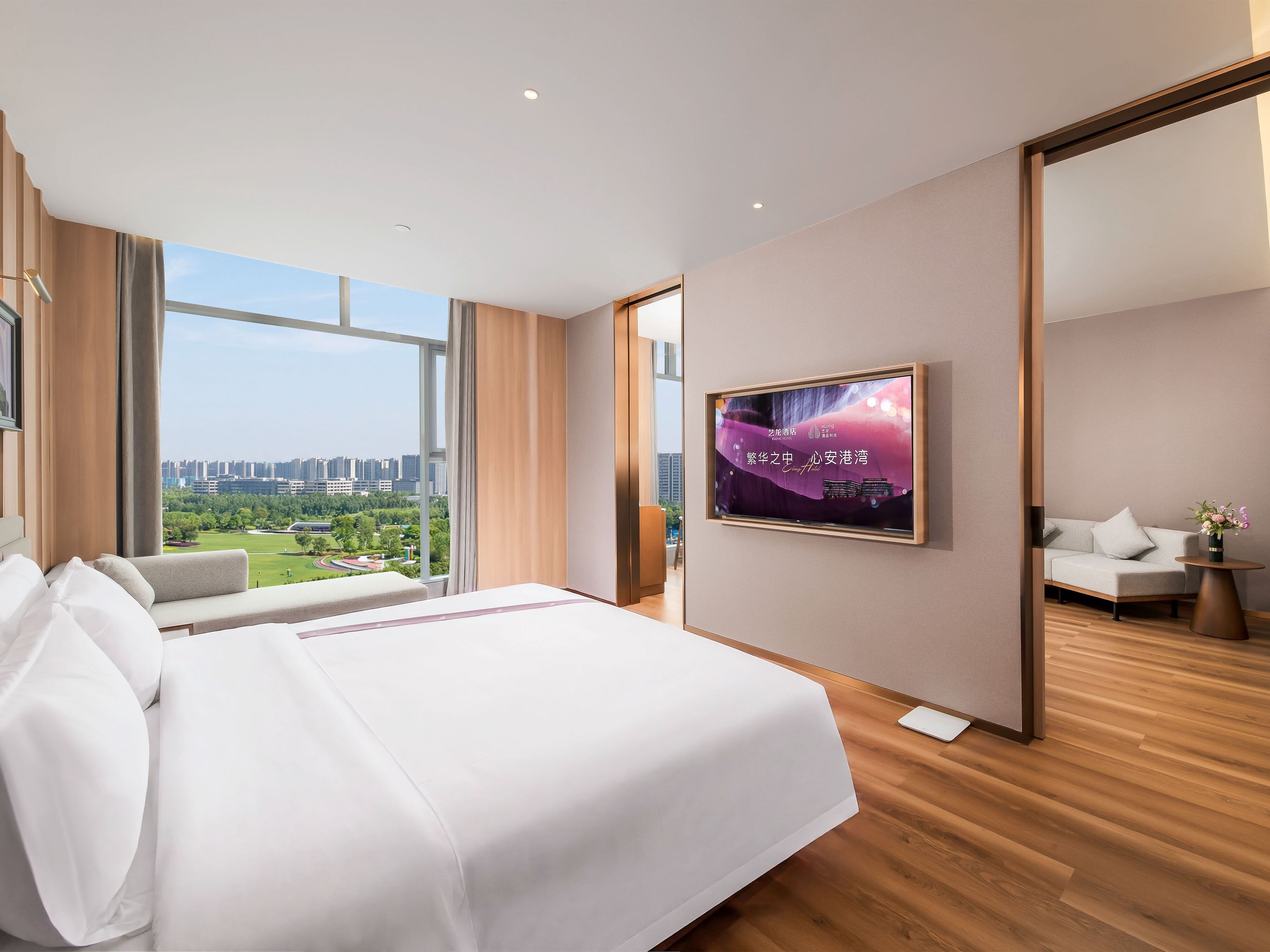A hotel room with a large bed, a TV mounted on the wall, and a view of a cityscape through large windows.