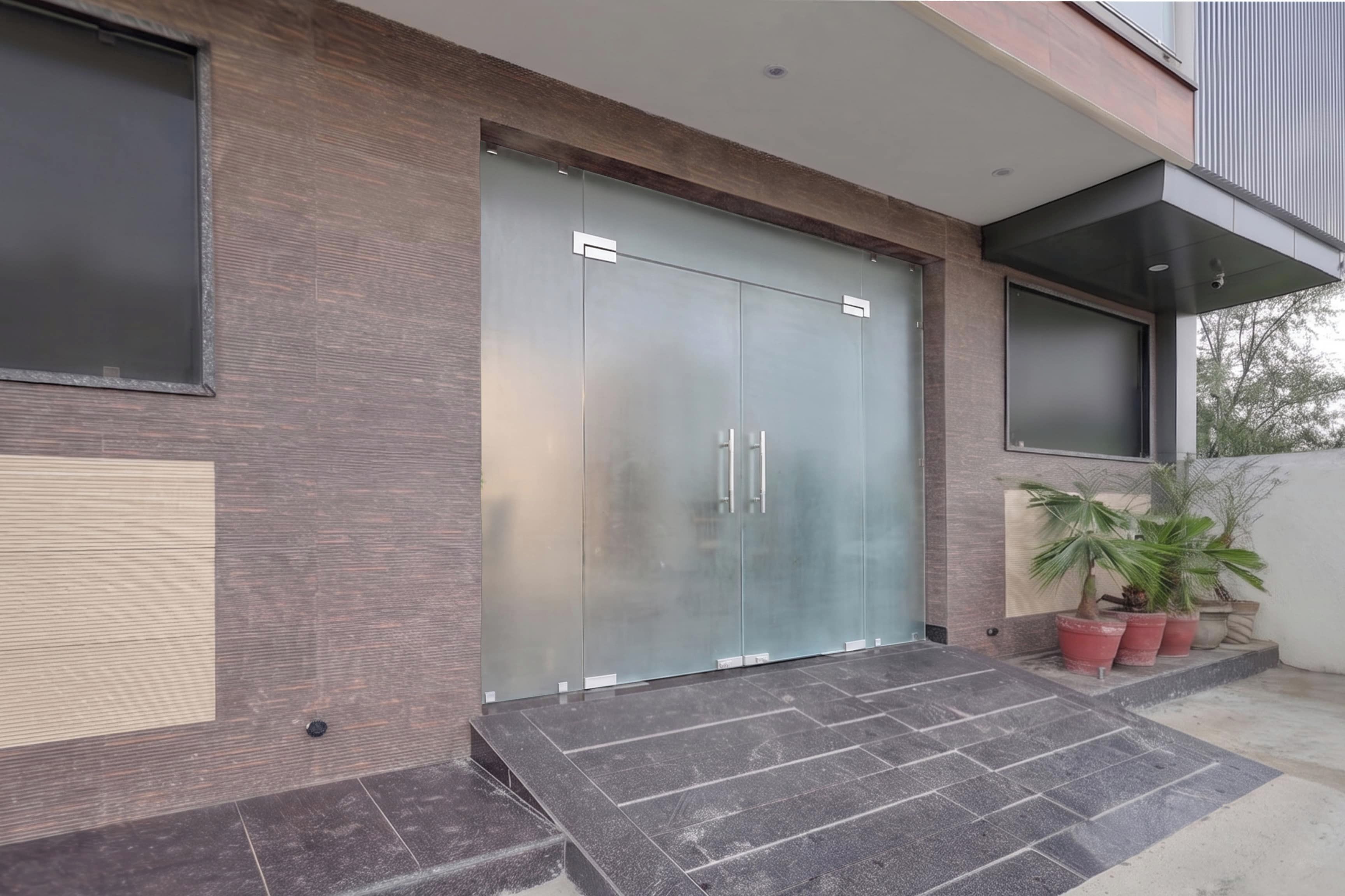 A modern building entrance with a glass door, a small outdoor area with potted plants, and a tiled floor.