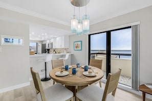 A dining area with a round table, chairs, and blue mugs. There is a kitchen in the background with white cabinets and a view of the sea through large windows. - Beachfront Bliss with Coastal Charm & Island Views - Condo Islands End 601 - Roelens (Fort Myers Beach)