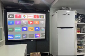 Ein Zimmer mit einem großen Bildschirm, der verschiedene App-Symbole anzeigt, einem weißen Kühlschrank und einem Tisch mit einem Stuhl.