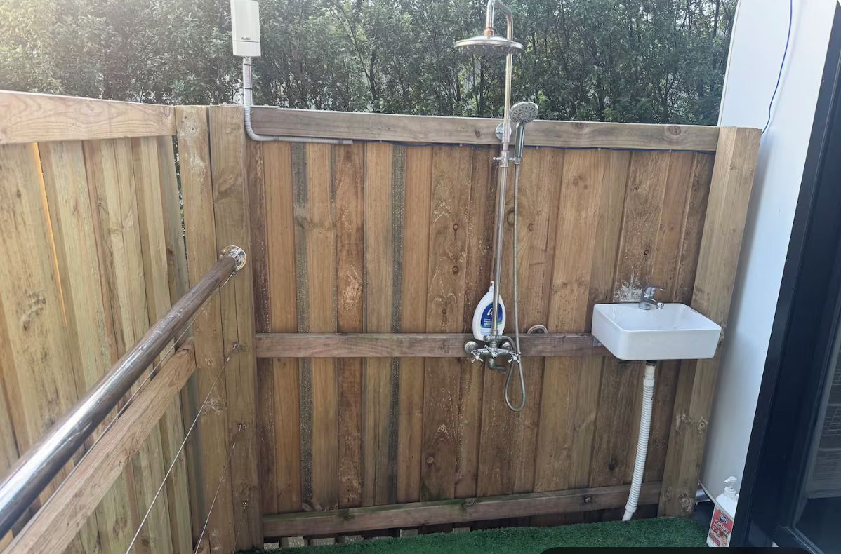 Un espace clos délimité par une clôture en bois, comprenant une douche, un lavabo et une main courante.