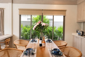 A dining area with a wooden table set for four, surrounded by wicker chairs, with a large window offering a view of greenery outside. - Stylish 2 Bedrooms Villa with Pool & Tropical Garden (Tambon Rawai)