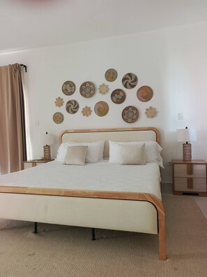 A neatly made bed with a wooden frame and white bedding, a wall with decorative woven art, a bedside table with a lamp, and a curtain on the window. - Paradise Private Pool Villa (Punta Cana)