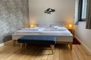 Un lit bien fait, avec du linge de lit blanc, un banc bleu et un motif décoratif représentant un paysage de montagne sur le mur.