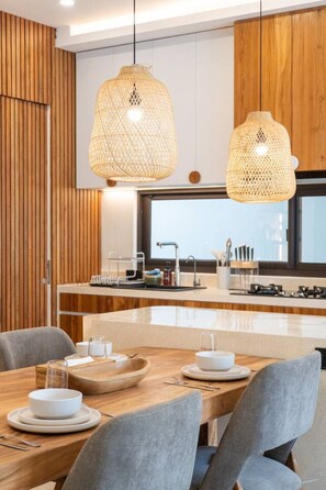 A modern kitchen with wooden cabinetry, a central island, pendant lights, and a dining area with grey chairs. - 2BR Batu Bolong Luxury Smart Mediterranean Villa (Kuta Utara)