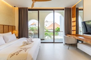 Ein Hotelzimmer mit Bett, Schreibtisch, Fernseher und einem Balkon mit Blick auf Gebäude und Grünflächen.