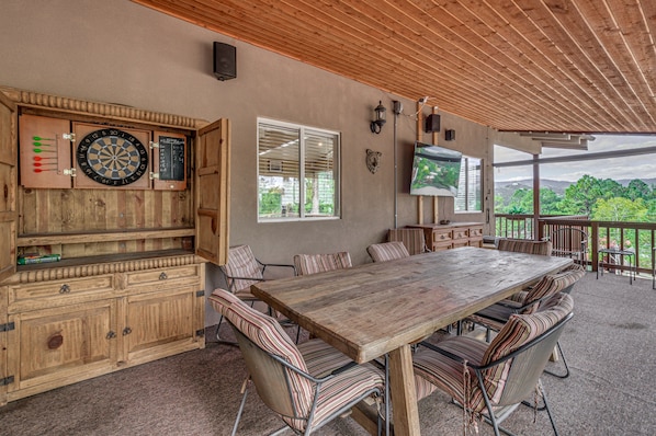 A covered outdoor dining area with a wooden table and chairs, a dartboard, and a television mounted on the wall. - Walk to Midtown! Mountain-View Ruidoso Retreat (Ruidoso)