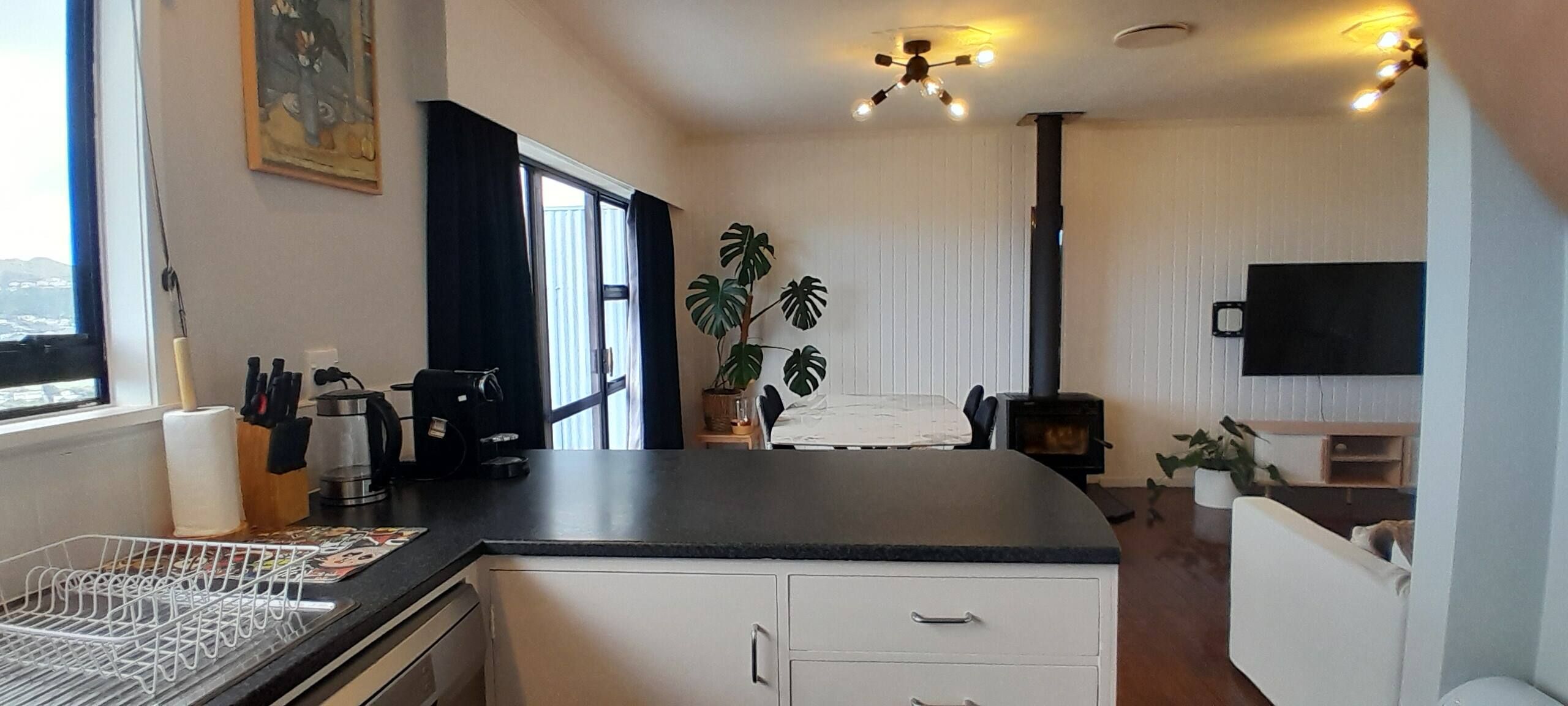 A compact kitchen with a coffee machine, a dining area with a table and chairs, a potted plant, and a TV mounted on the wall.