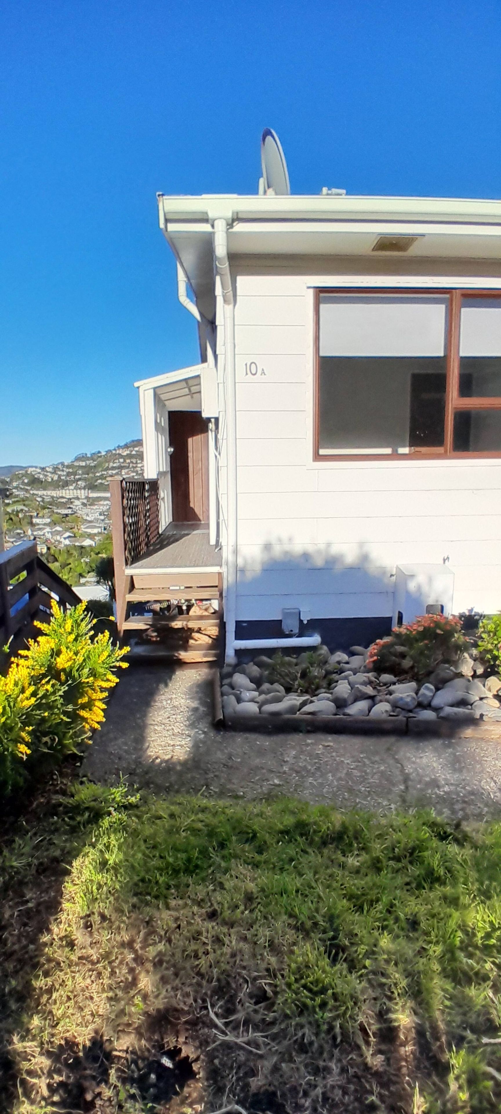 A single-story house with a visible number '10A' on the front wall, a satellite dish on the roof, and a small garden area with rocks and plants.