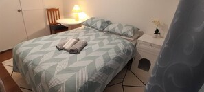 Un letto rifatto con cura, una coperta a motivi decorativi, un comodino con una lampada e un tavolino con una pianta.