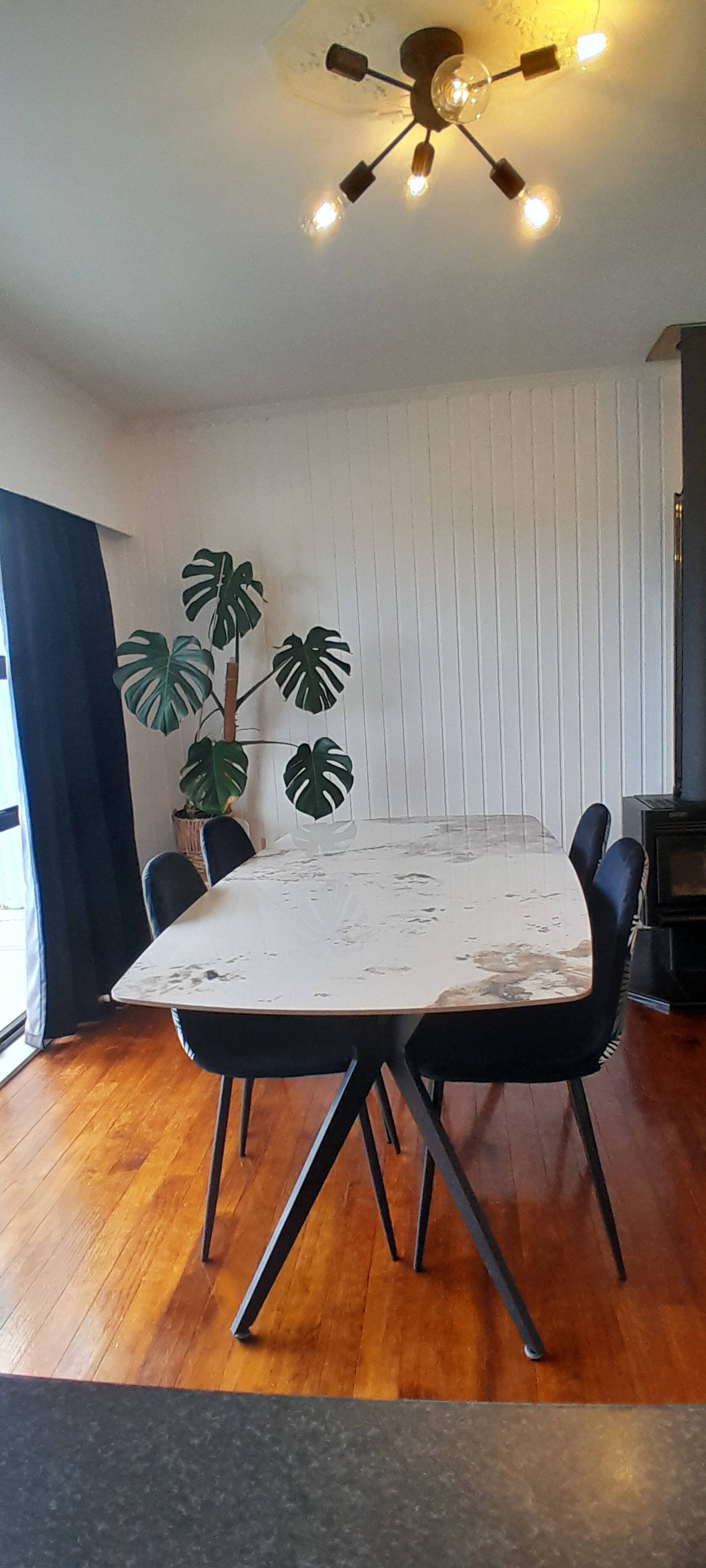 Zona pranzo con tavolo bianco, sedie nere, lampadario a sospensione e una pianta in vaso.
