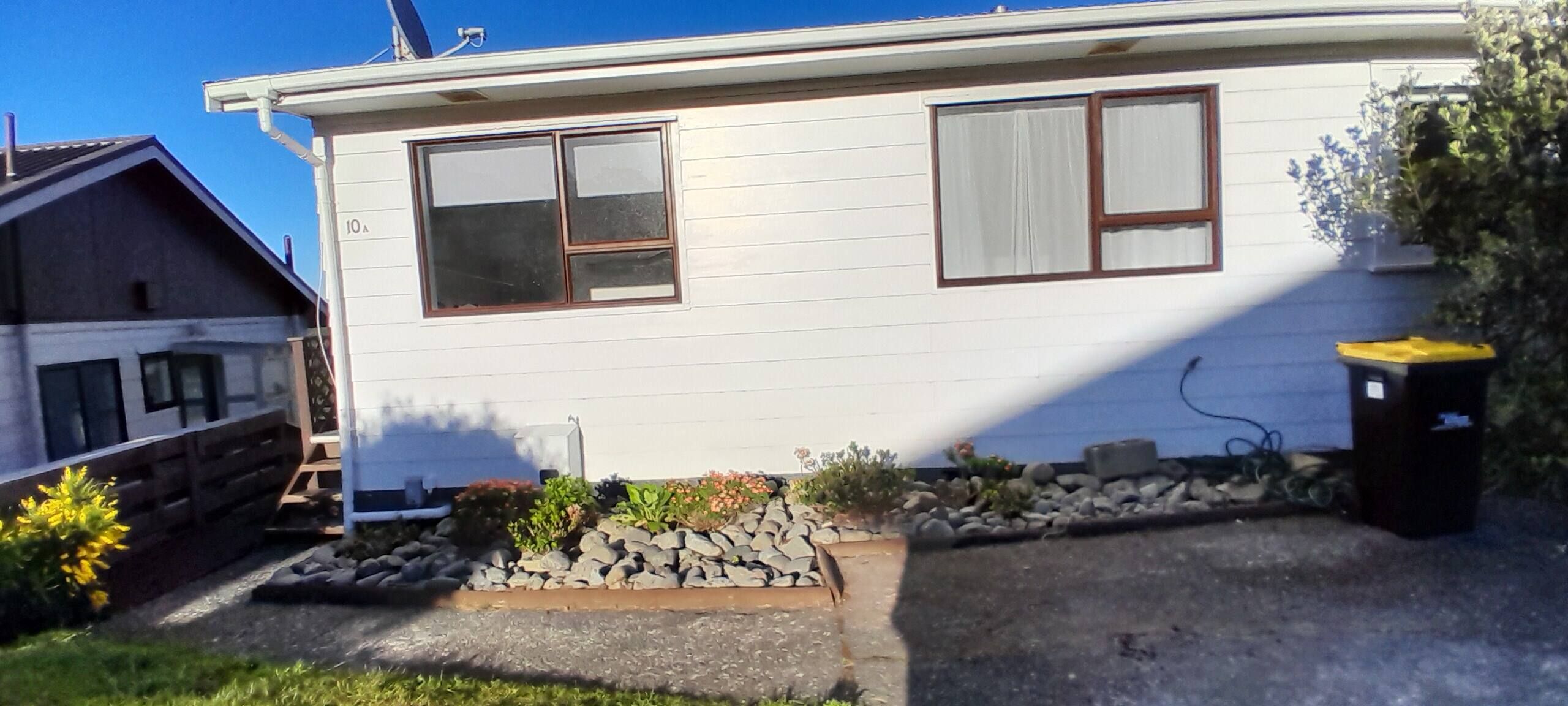 A single-story house with a front porch, a small garden with rocks and plants, and a trash can.