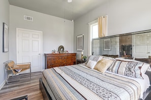 A bedroom with a large bed, a wooden dresser, a chair, and a mirror. - New | Veteran Host | Luxe | Book Now for Wow (Galveston)