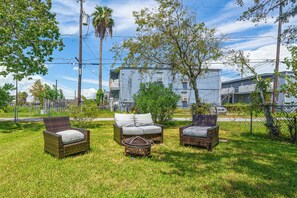 An outdoor seating area with wicker furniture and a metal table, surrounded by grass and trees, with buildings in the background. - New | Veteran Host | Luxe | Book Now for Wow (Galveston)