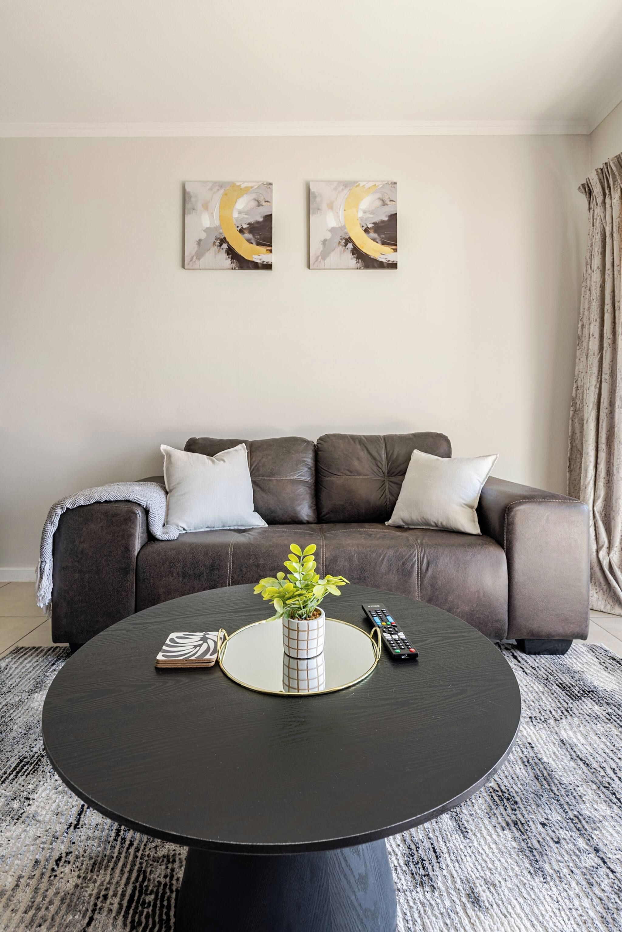 Una sala de estar con un sofá de cuero marrón, dos almohadas blancas, una mesa de centro redonda negra con una planta, un control remoto y una revista.