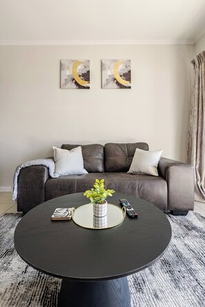 A living room with a brown leather sofa, two white pillows, a round black coffee table with a plant, a remote control, and a magazine. - Beautiful view with Netflix and Fast Wifi (Randburg)