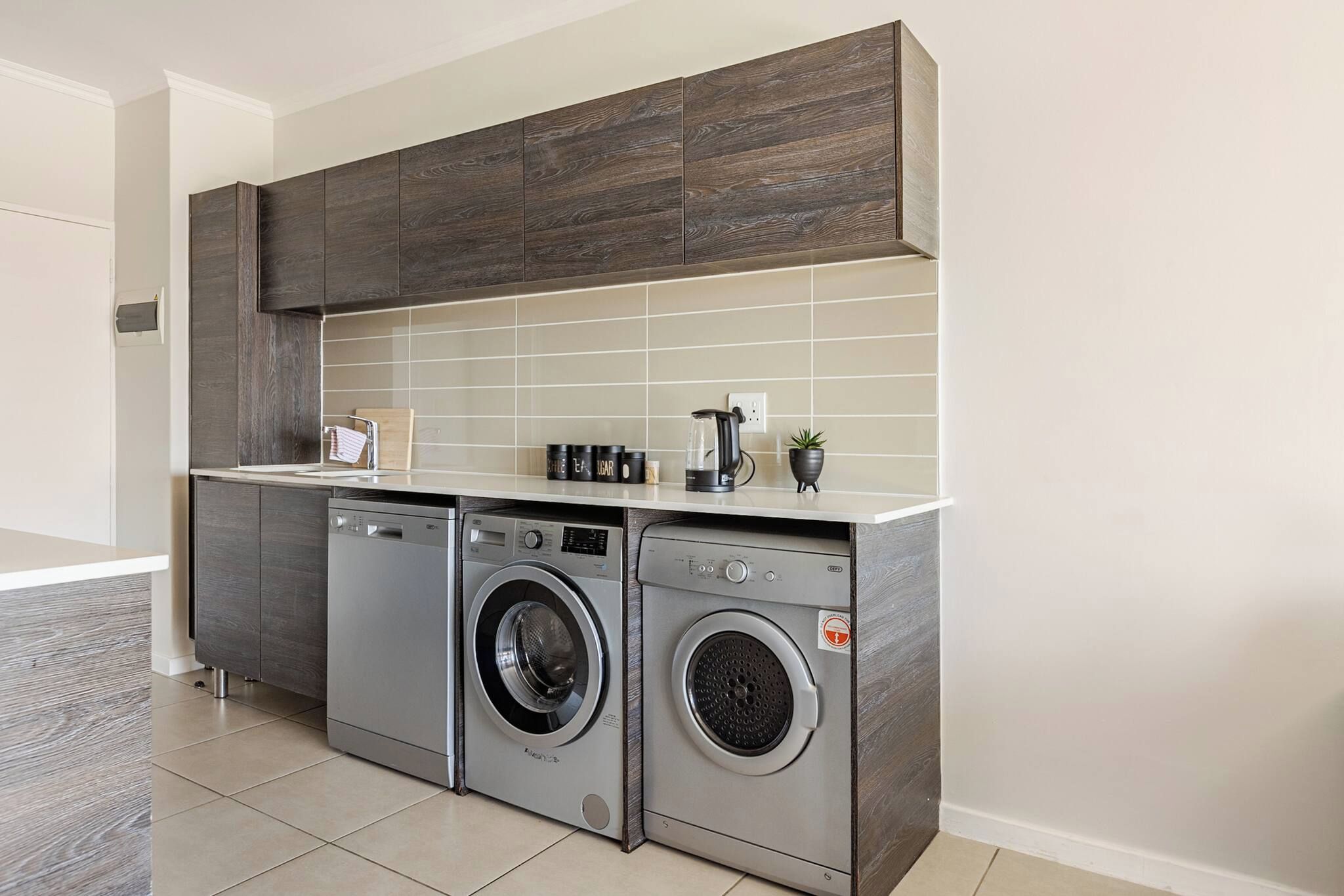 A compact laundry area with two front-loading washing machines and a dryer, a small countertop with a kettle and a plant, and a tiled backsplash.