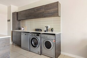 A compact laundry area with two front-loading washing machines and a dryer, a small countertop with a kettle and a plant, and a tiled backsplash. - Beautiful view with Netflix and Fast Wifi (Randburg)