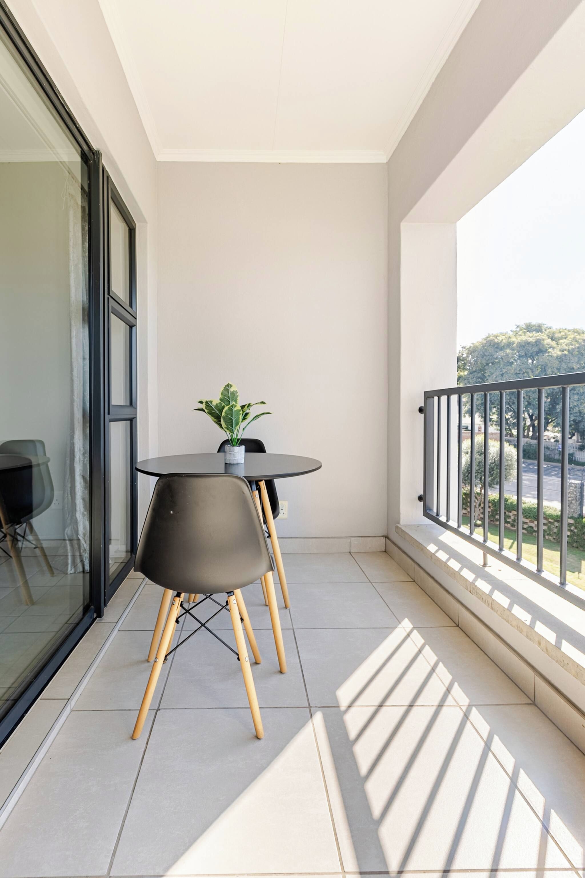 A balcony with a round table and two chairs, a potted plant, and a view of the street.