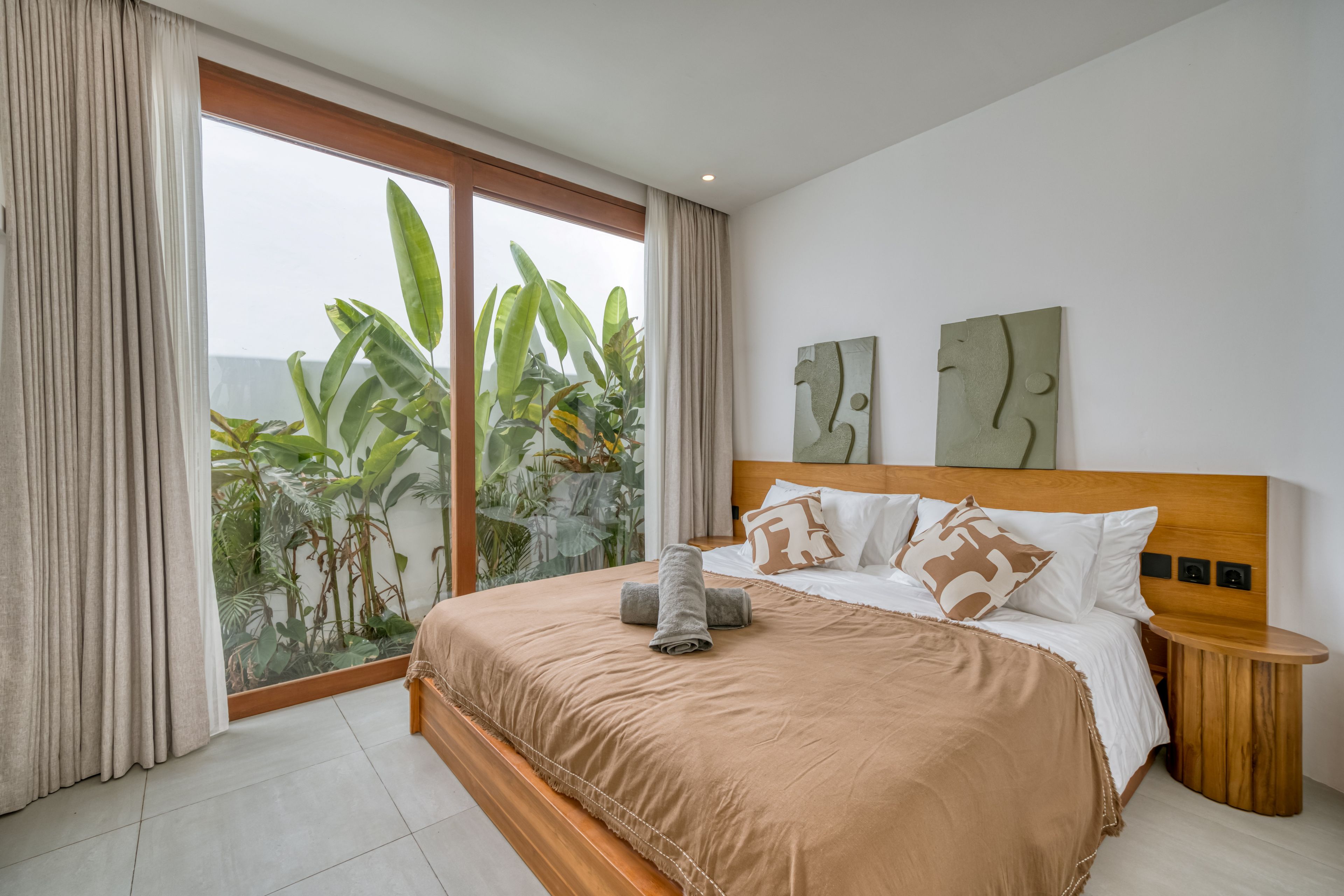 Une chambre moderne avec un grand lit, une tête de lit en bois et une vue sur des plantes tropicales, visible à travers une porte-fenêtre coulissante.