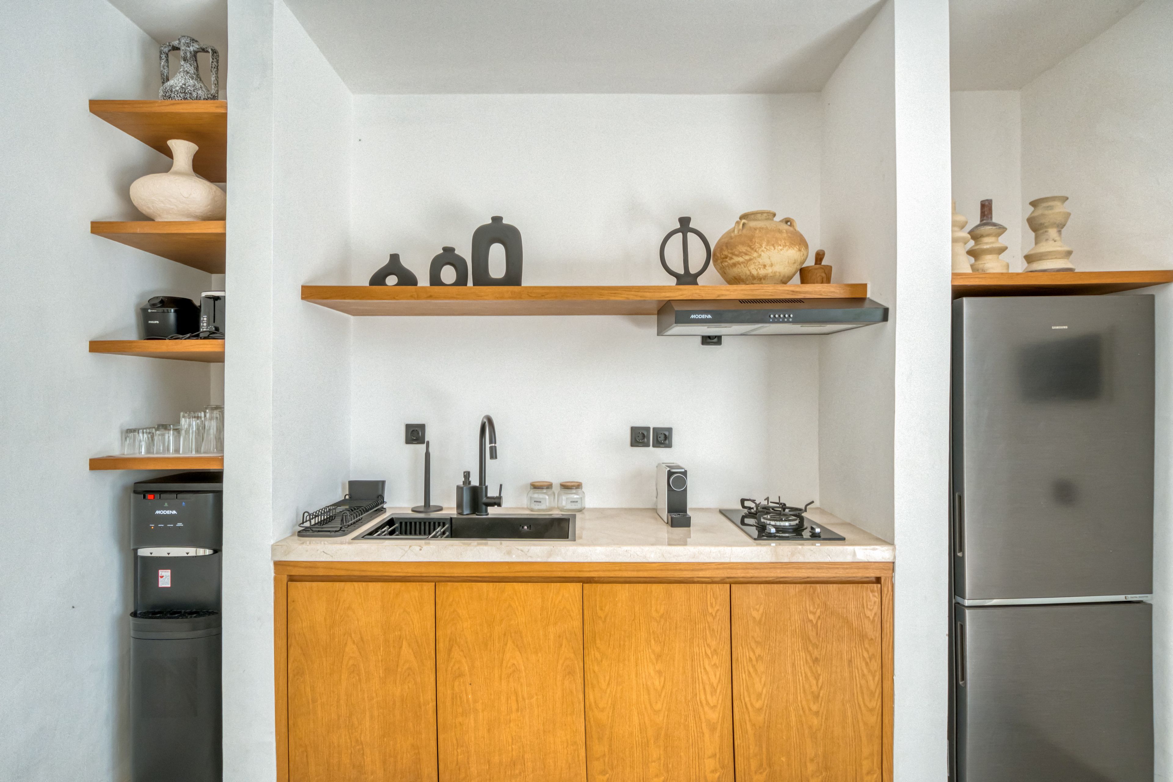 Une cuisine moderne avec des meubles en bois, un réfrigérateur en acier inoxydable, un four encastré, une machine à café et des objets décoratifs disposés sur des étagères.