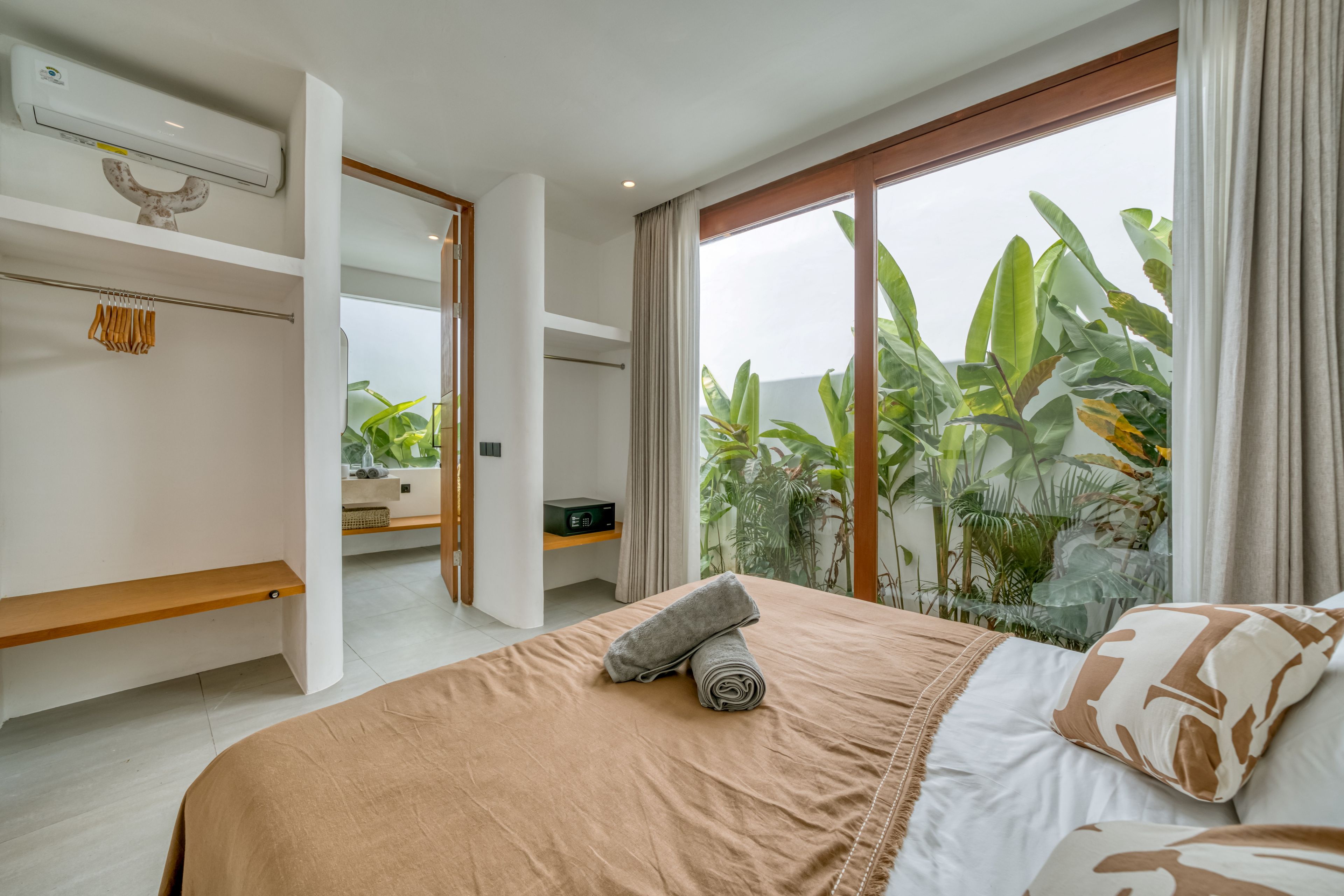 A modern bedroom with a large bed, a wooden shelf, and a view of tropical plants through the window.