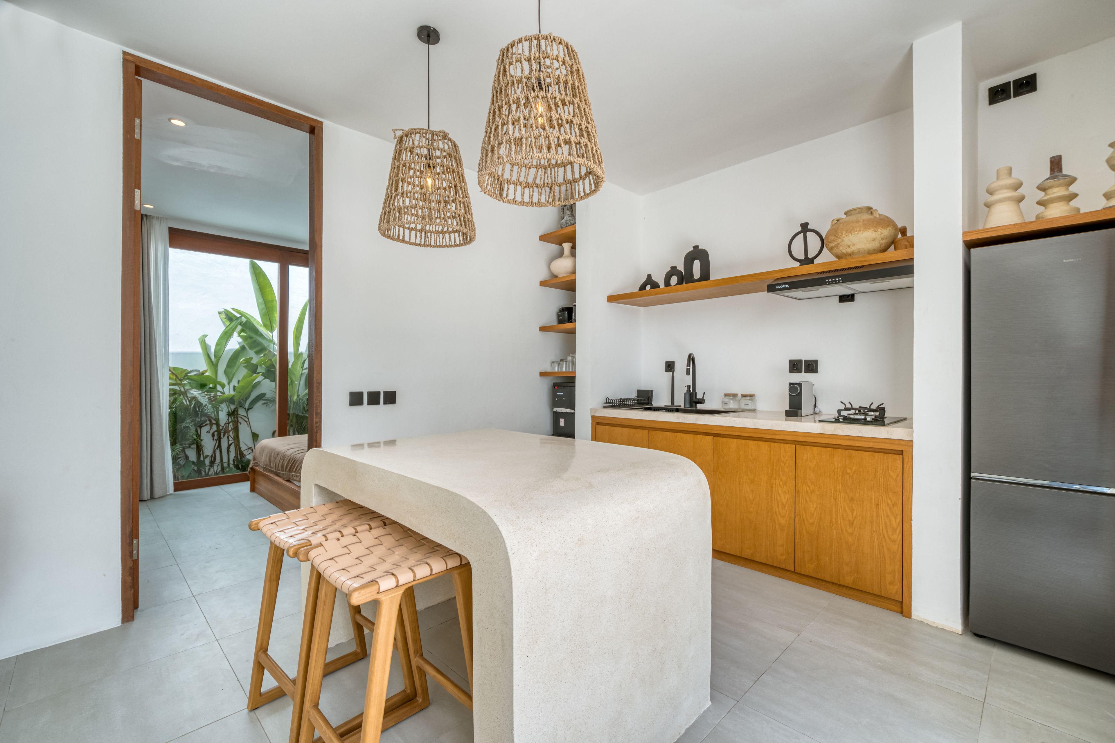 A modern kitchen with a central island, wooden stools, hanging wicker light fixtures, and a stainless steel refrigerator.