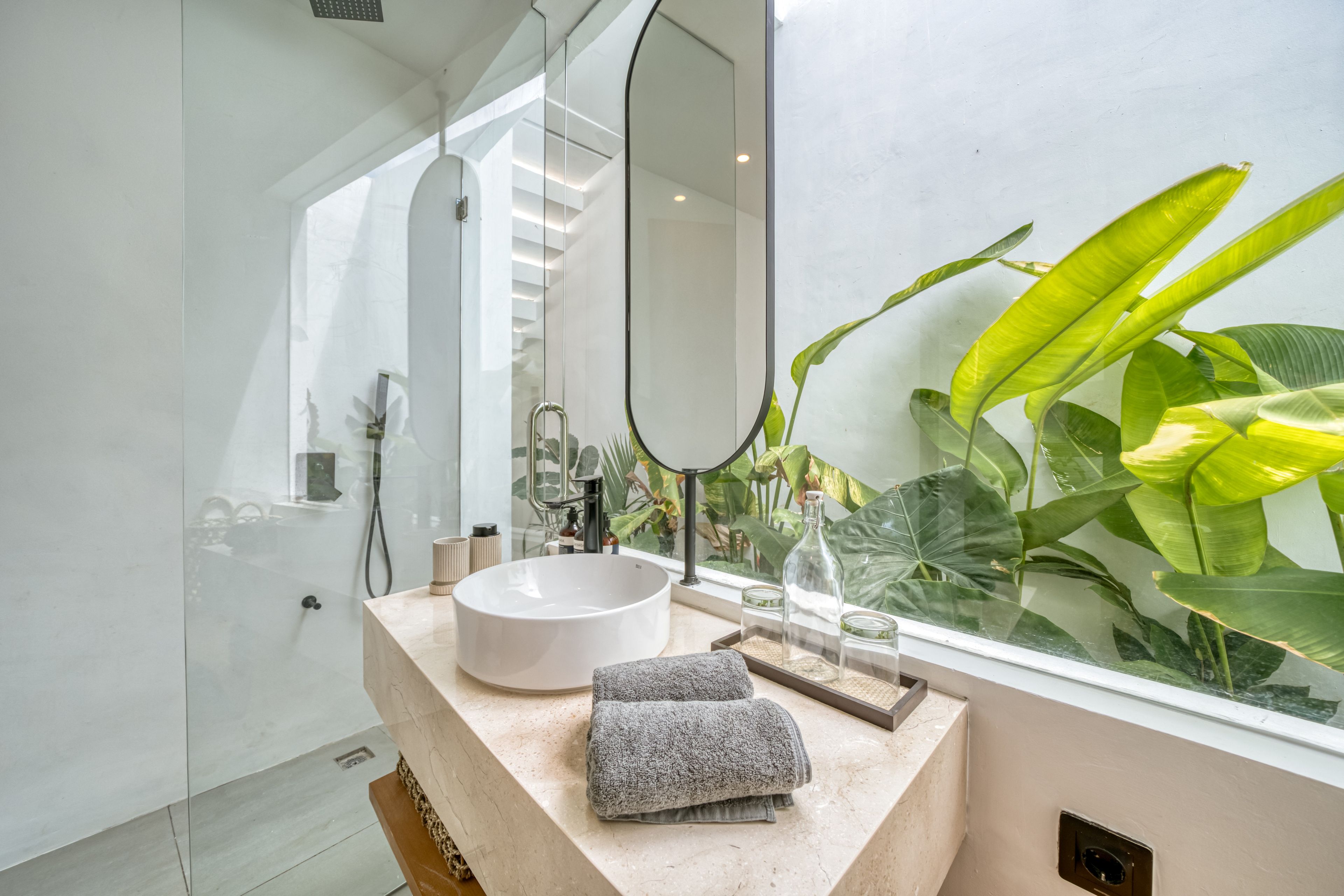 A modern bathroom with a large mirror, a round sink, and a window with tropical plants.