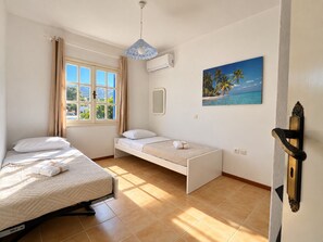 Una habitación con cama, mesita de noche, ventana con cortinas, equipo de aire acondicionado, un cuadro de playa y una puerta con manija.