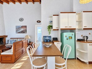 A cozy kitchen with a wooden dining table, white chairs, a teal refrigerator, and a view of the sea through the window. - Artemis Sea Cottage - Near beach & airport by ΜΡS (Artemis)
