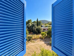Vista a través de contraventanas azules hacia un jardín con árboles y un edificio al fondo.