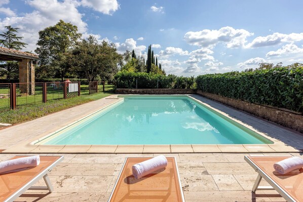 Una piscina rectangular con superficie de agua azul cristalina, rodeada por un muro de piedra bajo y una cerca metálica. Hay sillas de descanso con toallas sobre ellas, y el área está delimitada por un seto.