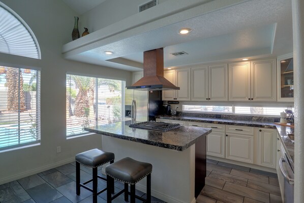 A kitchen with a central island, bar stools, white cabinets, and a view of the outdoors through large windows. - Private Pool & Patio: Modern Henderson Home (Henderson)